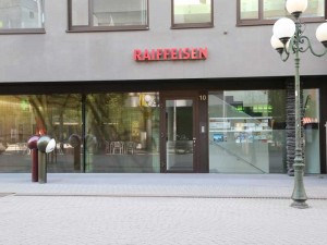 Eingang einer Raiffeisen Bankfiliale mit moderner Glasfassade und Straßenlaternen im Vordergrund.